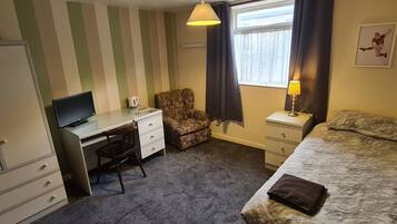 Habitación de hotel pequeña con una cama individual, un escritorio con silla, una mesita con lámpara y una ventana con cortinas.