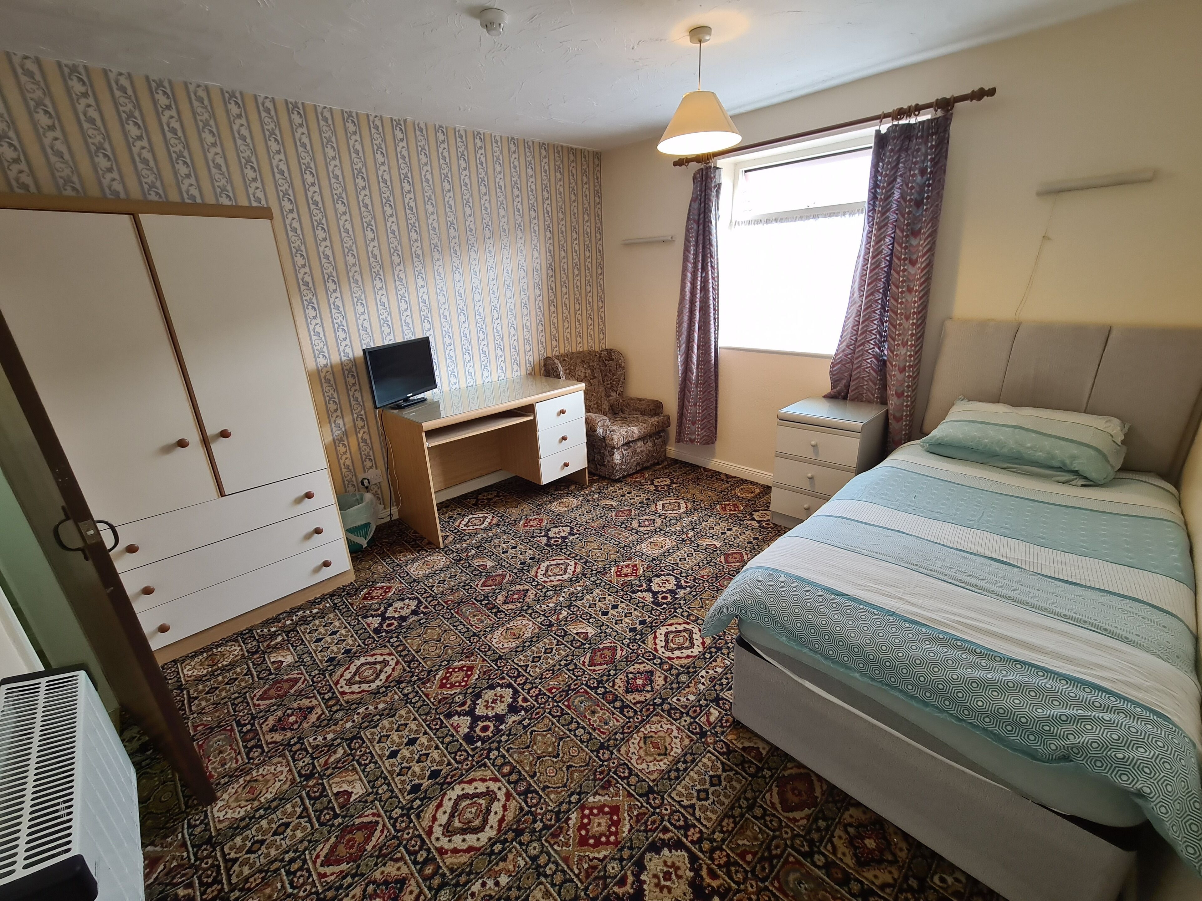 A bedroom with a bed, a desk, a chair, a wardrobe, patterned carpet, curtains, and a hanging light fixture.