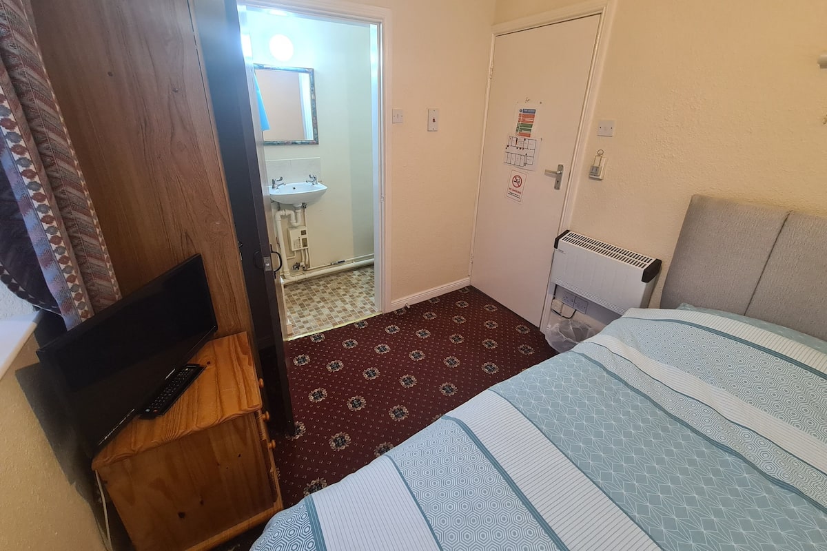 A bedroom with a bed, a wooden desk with a laptop, a radiator, and a bathroom visible through an open door.