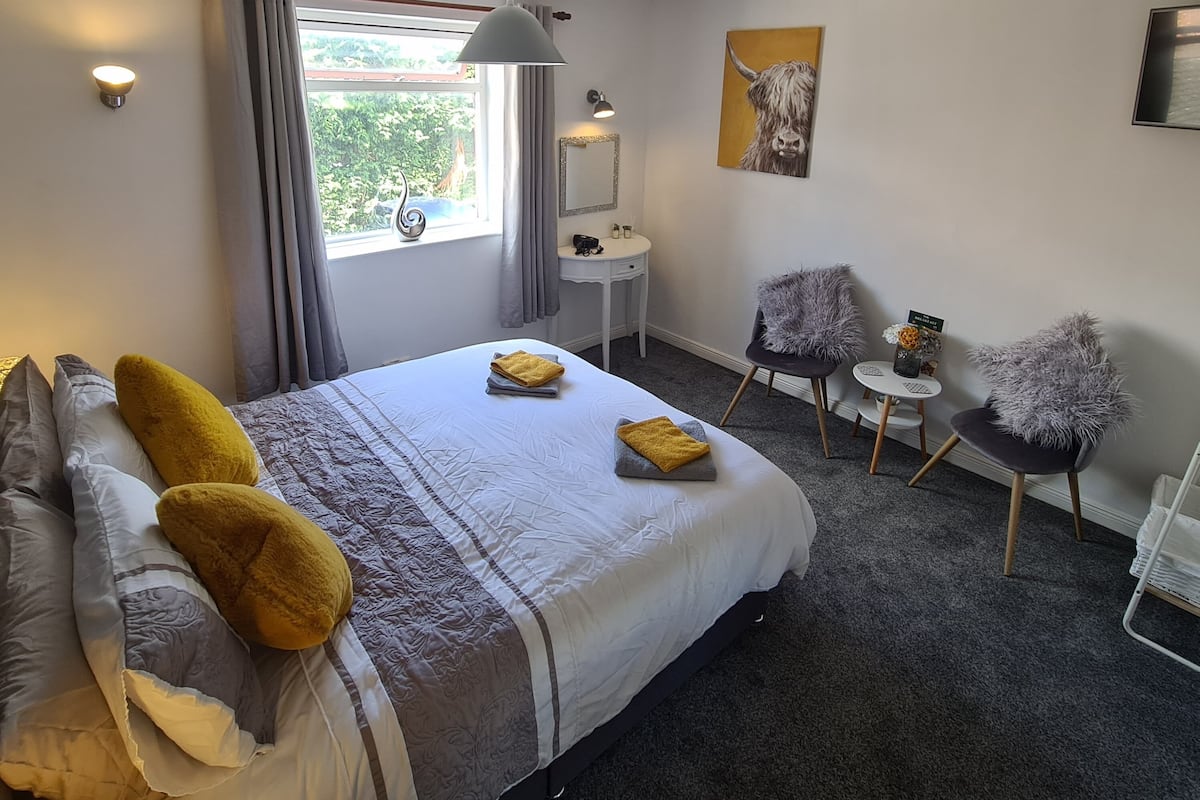 A hotel room with a bed, a desk, two chairs, a window with curtains, a painting, and a ceiling light.