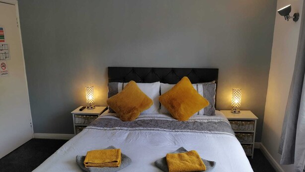 Una cama bien tendida con dos almohadas, una mesita de noche con una lámpara y un aplique de pared.