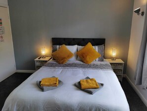 Una cama bien tendida con dos almohadas, una mesita de noche con una lámpara y un aplique de pared.