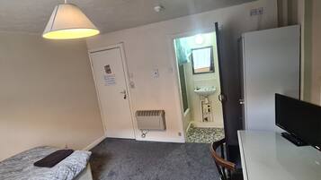 A small, sparsely furnished room with a bed, a desk with a computer, a chair, a TV, and a radiator.