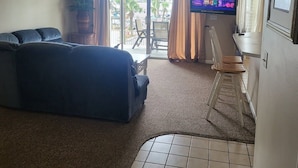 A living room with a sofa, a television, a ceiling fan, and a view of the outside.