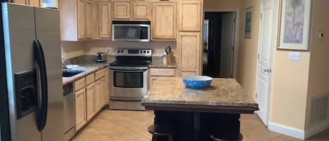 A kitchen with wooden cabinets, a stainless steel refrigerator, a microwave, an oven, and a granite countertop with a blue bowl.