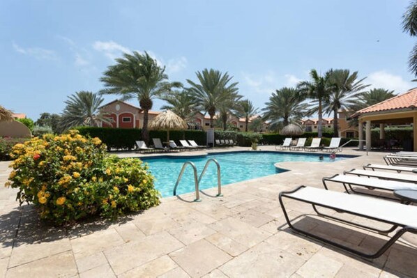 A pool area with lounge chairs, a swimming pool, palm trees, and a building with a red roof.