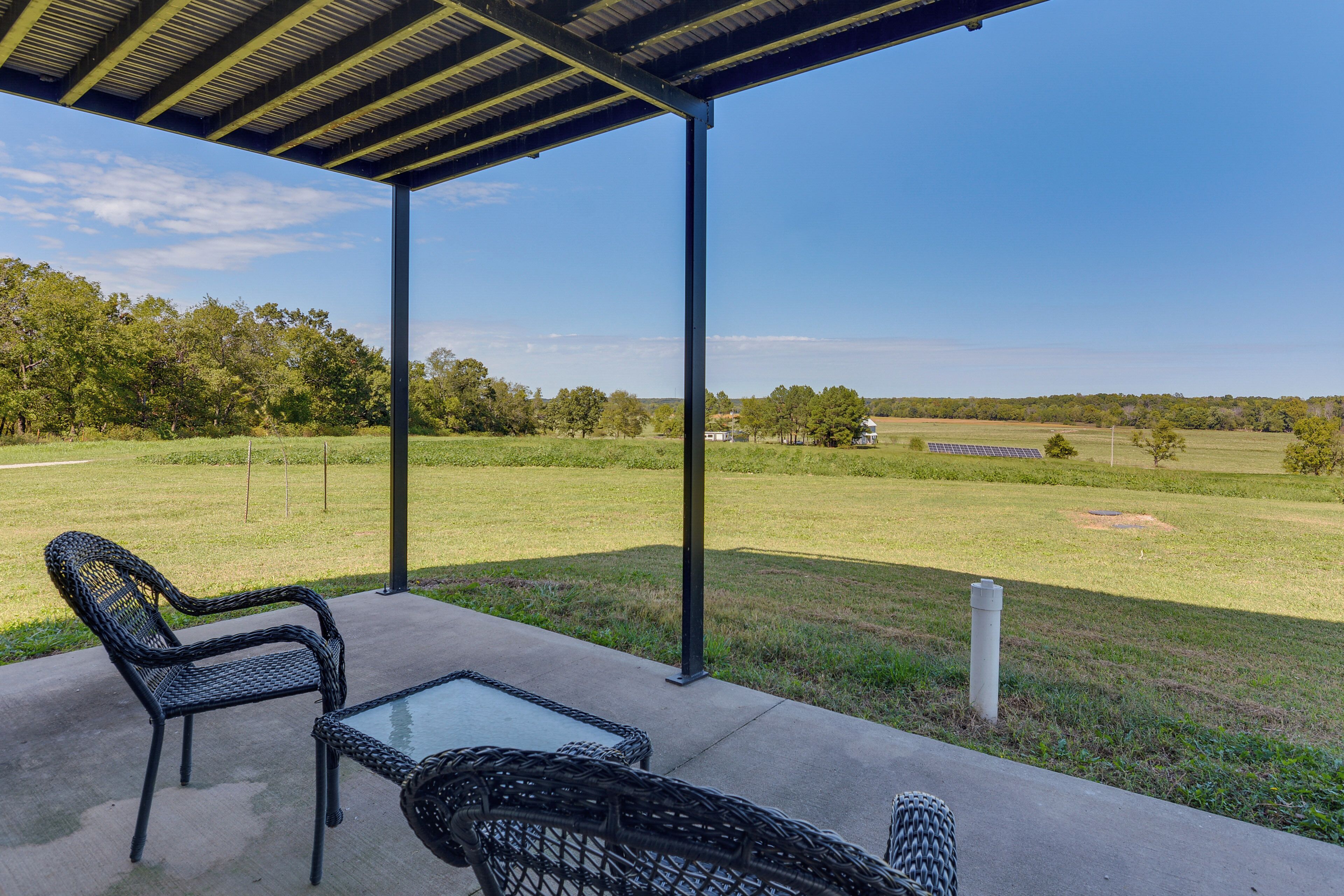 Área de varanda coberta com móveis de vime e mesa de vidro, com vista para um campo gramado e árvores ao longe.