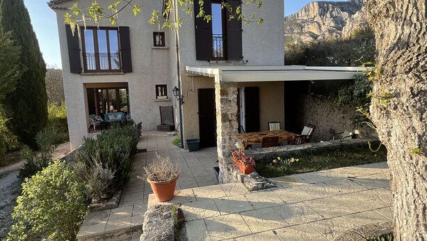 Une maison à deux étages avec une façade en pierre, un patio couvert et un jardin agrémenté de plantes en pot et d’un mur en pierre.