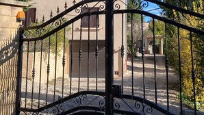 Une entrée fermée par un portail en fer forgé, un bâtiment à la façade beige et aux volets sombres, et un grand arbre à feuilles persistantes sur la droite.