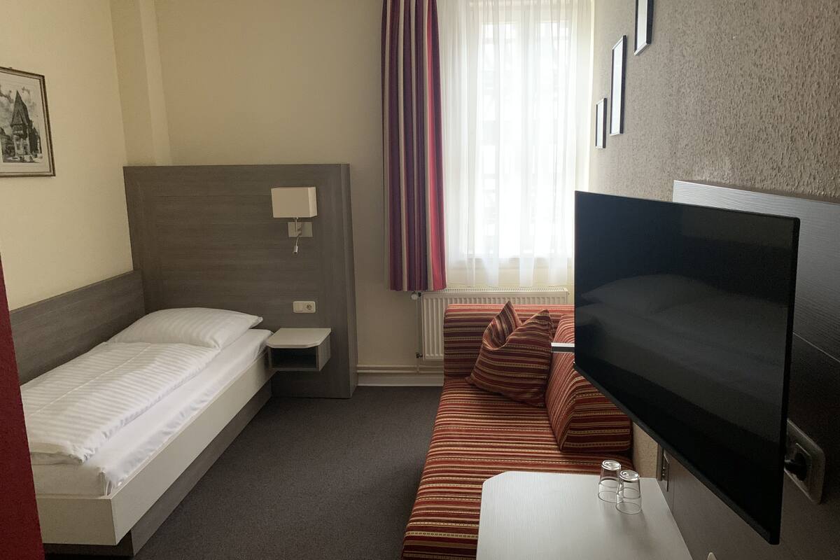 A hotel room with a single bed, a sofa, a TV, and a small table with glasses and a remote control.