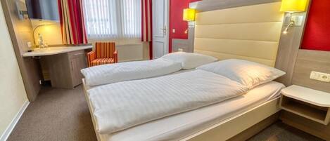 Habitación de hotel con cama doble, escritorio, silla, televisor y ventana con cortinas.