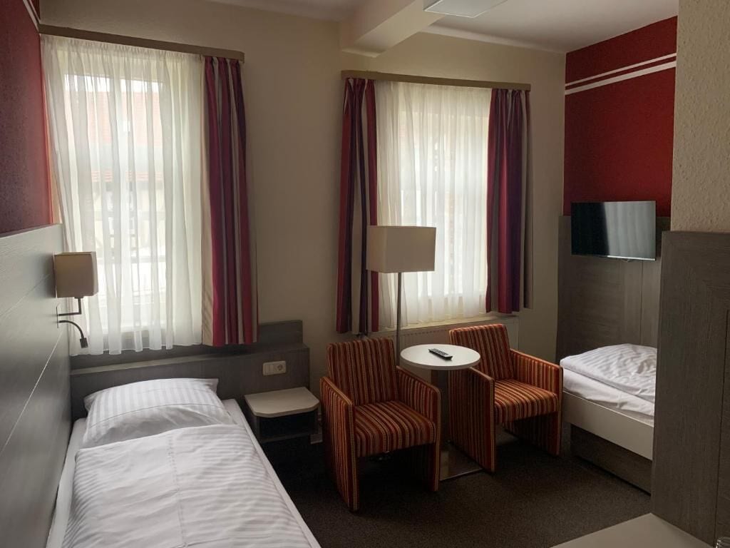Quarto de hotel com duas camas, uma mesinha com cadeiras, televisão e janela com cortinas.