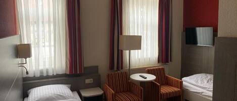 Una habitación de hotel con dos camas, una mesita con sillas, televisor y ventana con cortinas.