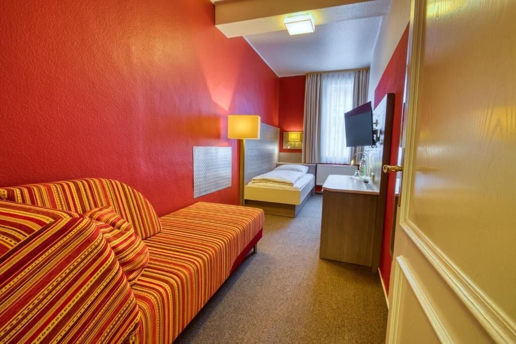 A hotel room with a red wall, a striped sofa, a bed, a desk with a lamp, and a television.