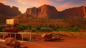 Un paysage désertique avec une charrette couverte posée sur une plateforme en bois, entourée de rochers et d’une végétation clairsemée.