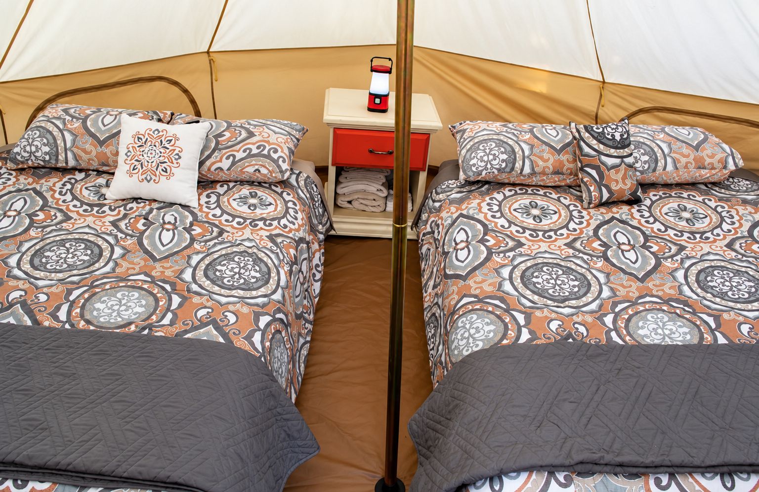 A tent with two beds, each with patterned bedding and pillows, a small table with a red drawer, and a white object on the table.