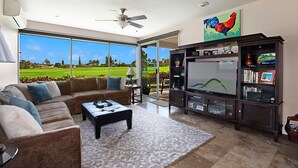 Un salón amplio con televisor grande, asientos cómodos y vistas a un campo de golf a través de amplias ventanas.
