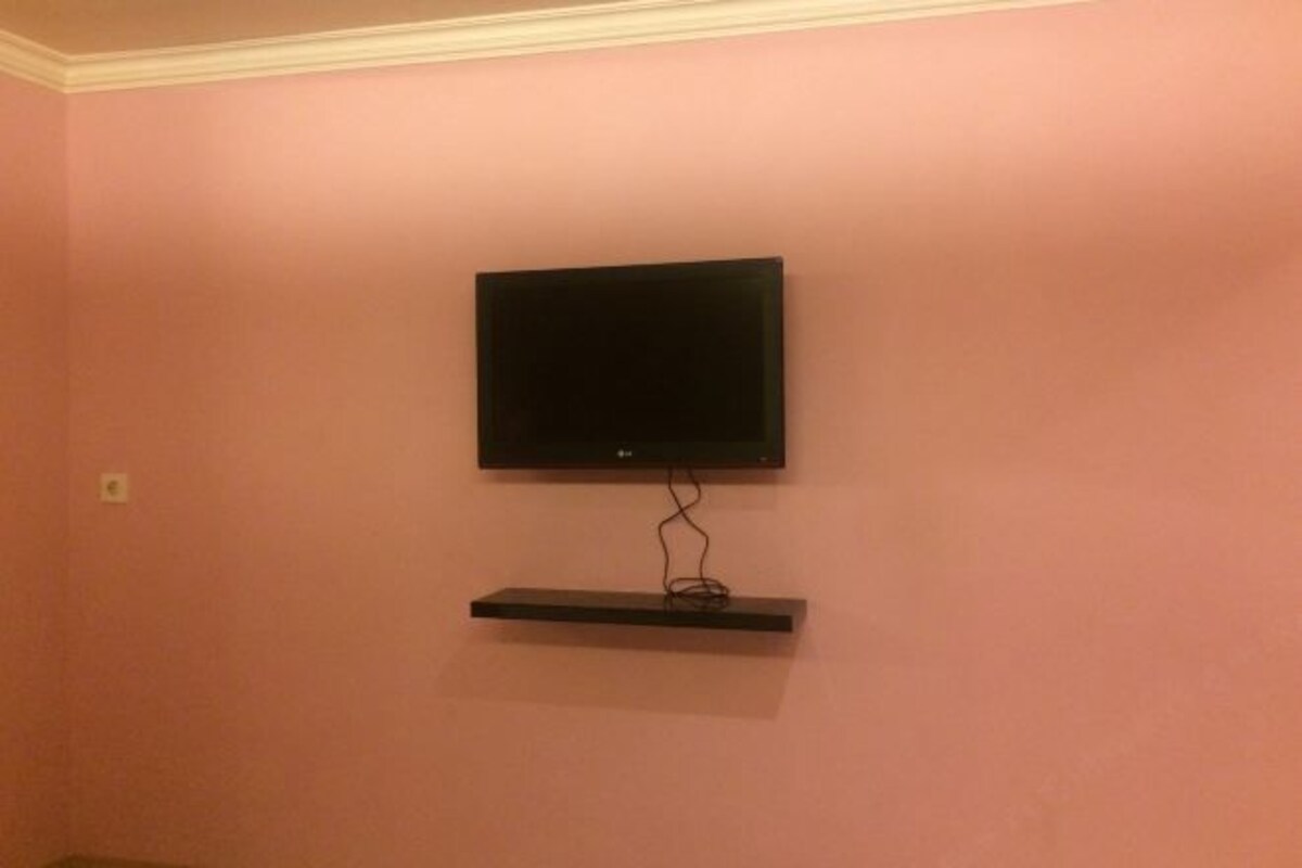 A television mounted on a pink wall with a black shelf below it.