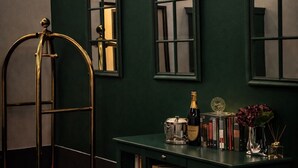 A dark green wall with the text "Sleepotels Suite Garde" and three windows. There is a metal cart with a red base, a bottle of champagne, and a small table with books and a vase of flowers. - Sleephotels Suite Garde (Hamburg)