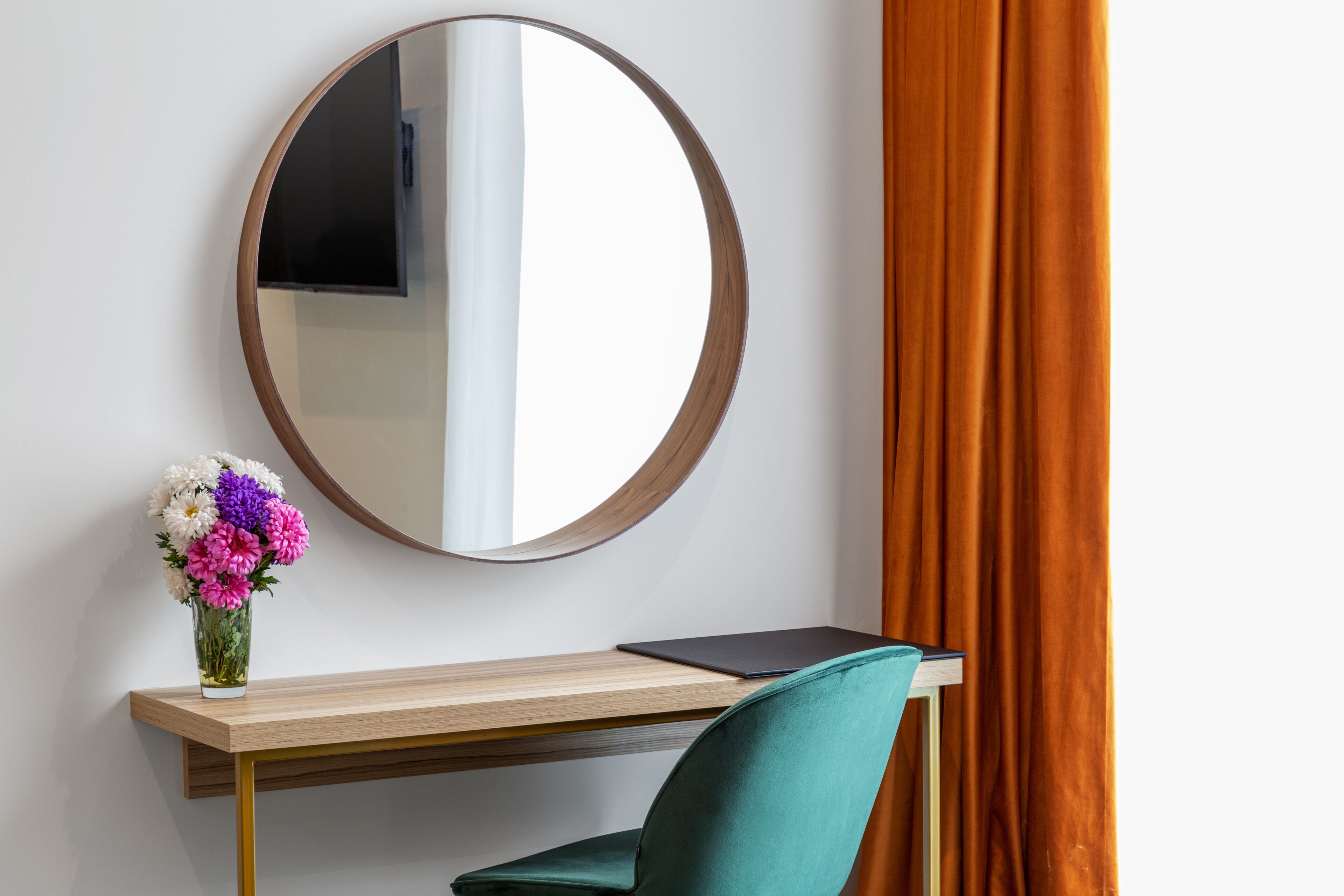 A desk with a laptop, a vase of flowers, and a round mirror on the wall.