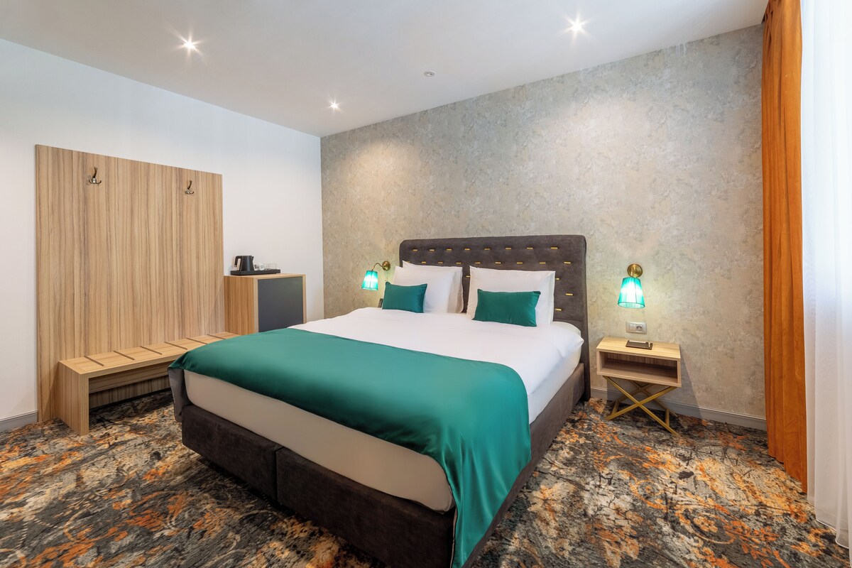 A hotel room with a large bed, a wooden wardrobe, a nightstand with a lamp, and a carpeted floor.