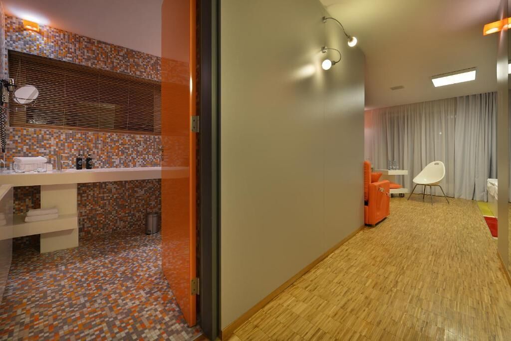 A modern bathroom with a tiled floor, a sink, and a mirror. There is a wooden floor in the adjacent room.