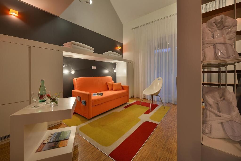 A modern hotel room with a sofa, a small table with a bottle and glasses, a chair, and a large window with curtains.