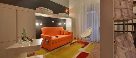 A modern hotel room with a sofa, a small table with a bottle and glasses, a chair, and a large window with curtains.