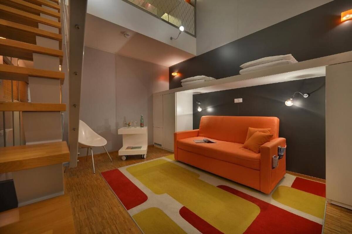 A modern living room with a wooden staircase, a bright orange sofa, and a colorful rug.