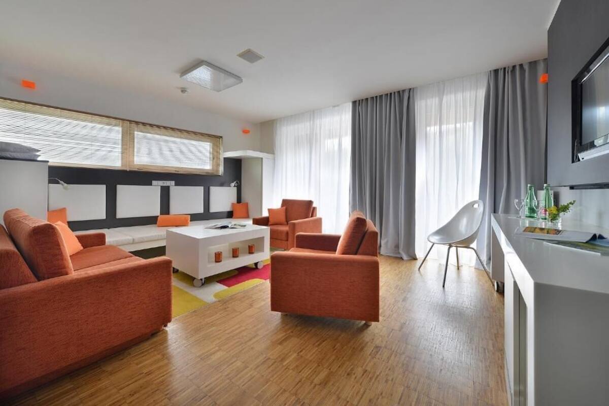 A modern living room with orange sofas, a white coffee table, and a kitchen area with a TV.