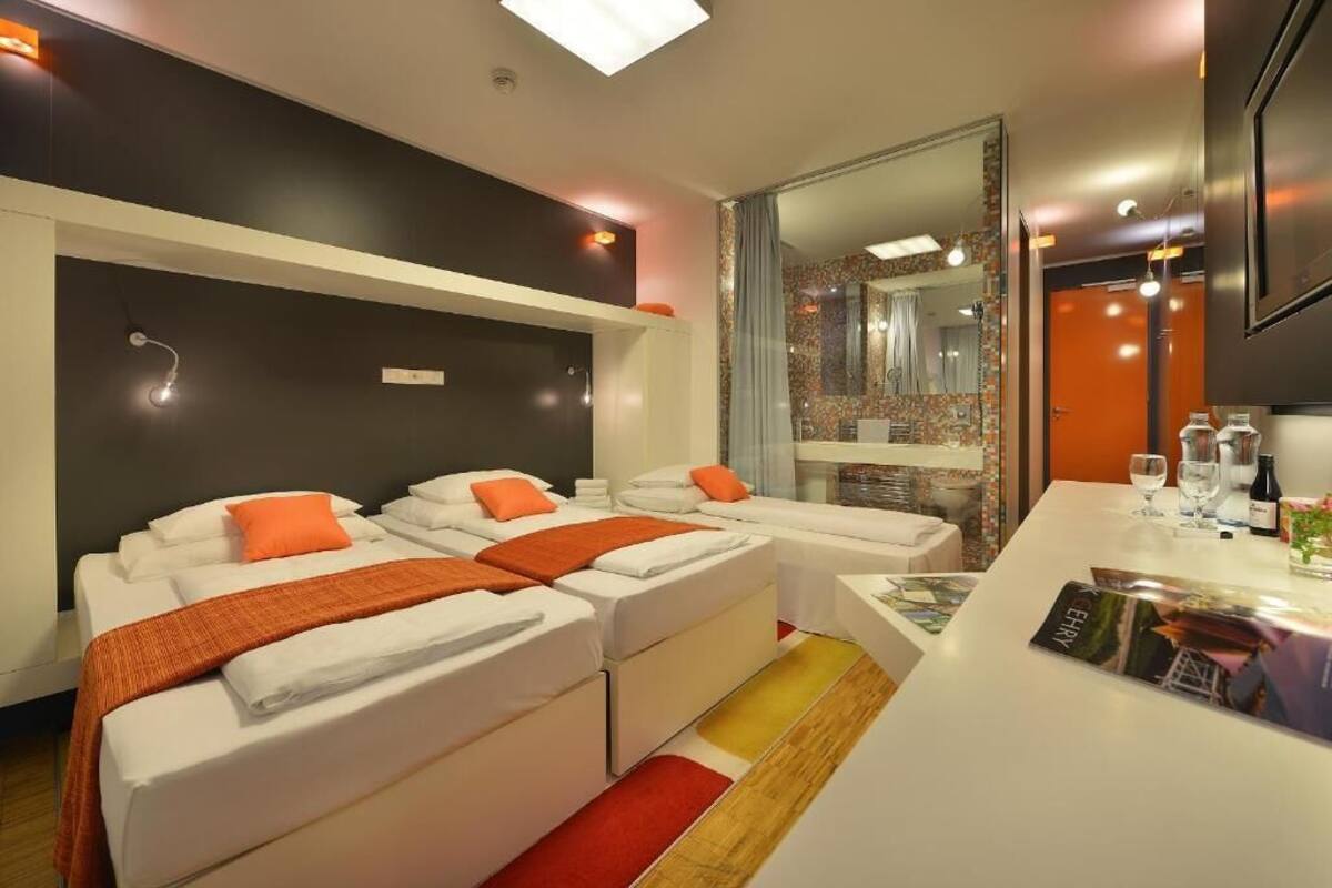 A modern hotel room with two beds, a small sitting area, a large mirror, and a minibar.