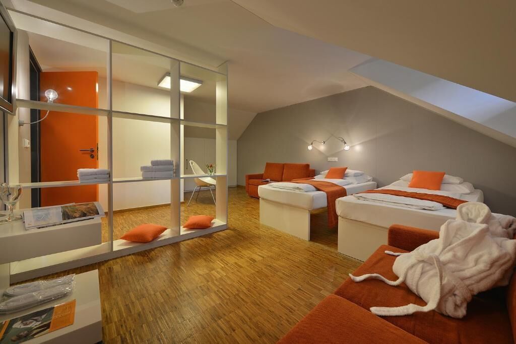 A modern hotel room with a loft-style ceiling, a glass partition, and a cozy seating area with orange cushions.