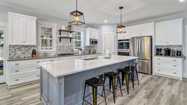 Cocina moderna con armarios blancos, una isla grande con taburetes de bar, electrodomésticos de acero inoxidable e iluminación colgante geométrica.