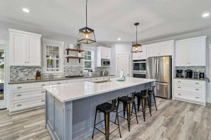 A modern kitchen with white cabinets, a large island with bar stools, stainless steel appliances, and geometric pendant lighting.