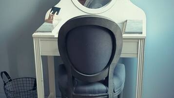 Een piano met daar boven een ronde spiegel, een zwarte stoel ervoor en een wit bureau met een mandje ernaast.