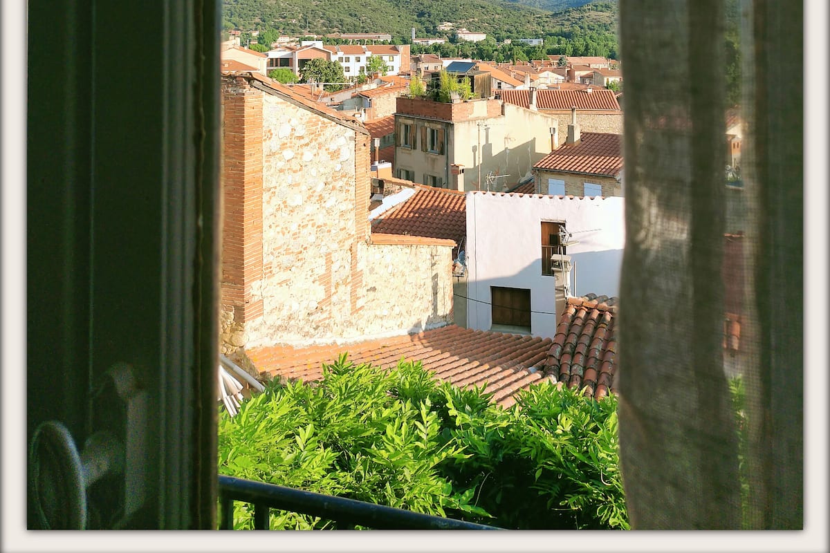 Vista desde una ventana que muestra un pueblo con edificios, vegetación y colinas a la distancia.
