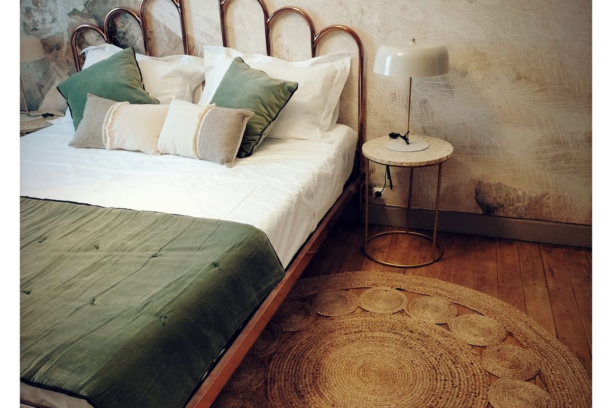 Una cama con colcha verde, varios almohadones, un tapete redondo tejido, una mesita de noche con lámpara y suelo de madera.