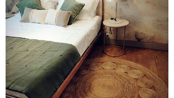 Un lit recouvert d’une couvre-lit verte, plusieurs oreillers, un tapis rond en fibres naturelles, une table de chevet avec une lampe et un plancher en bois.