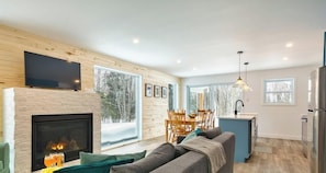 A modern living room with a fireplace, a television, and a dining area with a view of trees.