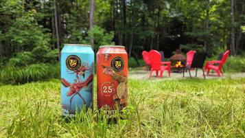 Two beverage cans on grass with a forest and outdoor seating in the background.