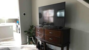 A living room with a flat-screen TV on a wooden cabinet, a kitchen in the background, and a potted plant. - Bluffton Retreat, Pet Friendly (Bluffton)