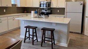 A modern kitchen with white cabinetry, a central island with stools, stainless steel appliances, and a window above the sink. - Bluffton Retreat, Pet Friendly (Bluffton)