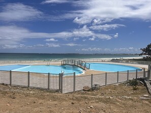 A pool area with two swimming pools, a beach, and a clear sky.