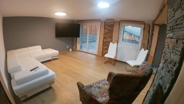 A living room with a white sofa, a floral armchair, a flat-screen TV mounted on the wall, and a large window with a view of a bridge. - Romantic Luxury appartment in the heart of Grachen (Grächen)