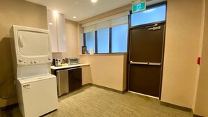 A compact hotel room with a kitchenette, a washer-dryer unit, a window with blinds, and an exit sign. - Room in luxurious downtown property (St. John's)