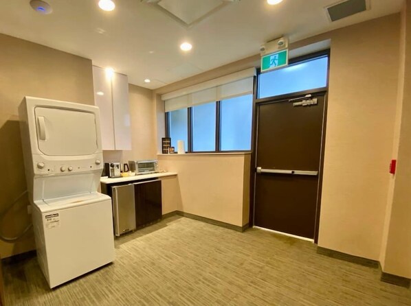 Habitación de hotel compacta con kitchenette, lavadora-secadora, ventana con persianas y señal de salida.