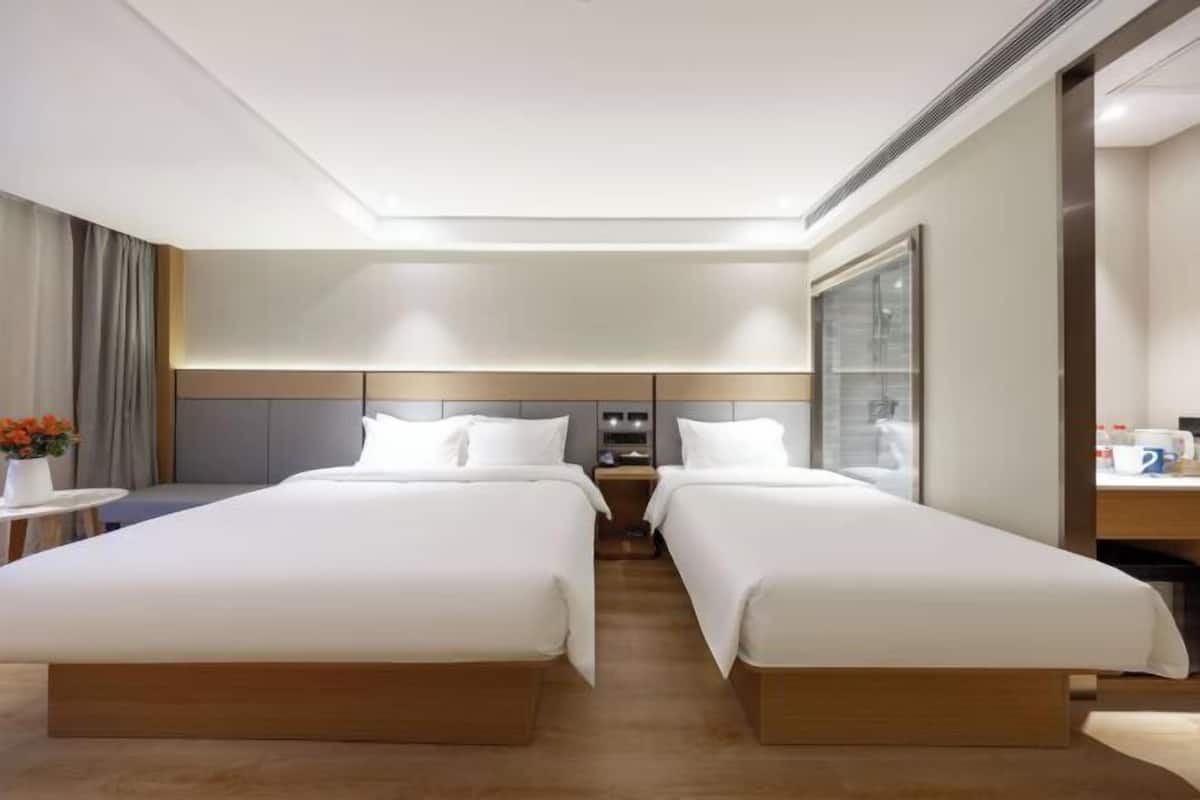 A hotel room with two beds, a small table with a vase of flowers, a nightstand with a lamp, and a large window.