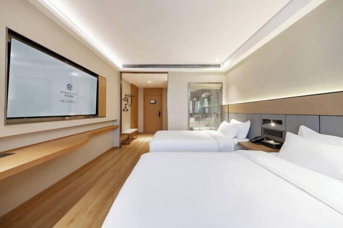 A modern hotel room with two beds, a large flat-screen TV, wooden flooring, and a view of the city through a glass wall.