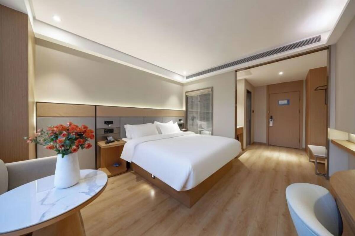 A modern hotel room with a large bed, a bedside table, a chair, a desk, and a vase with flowers.