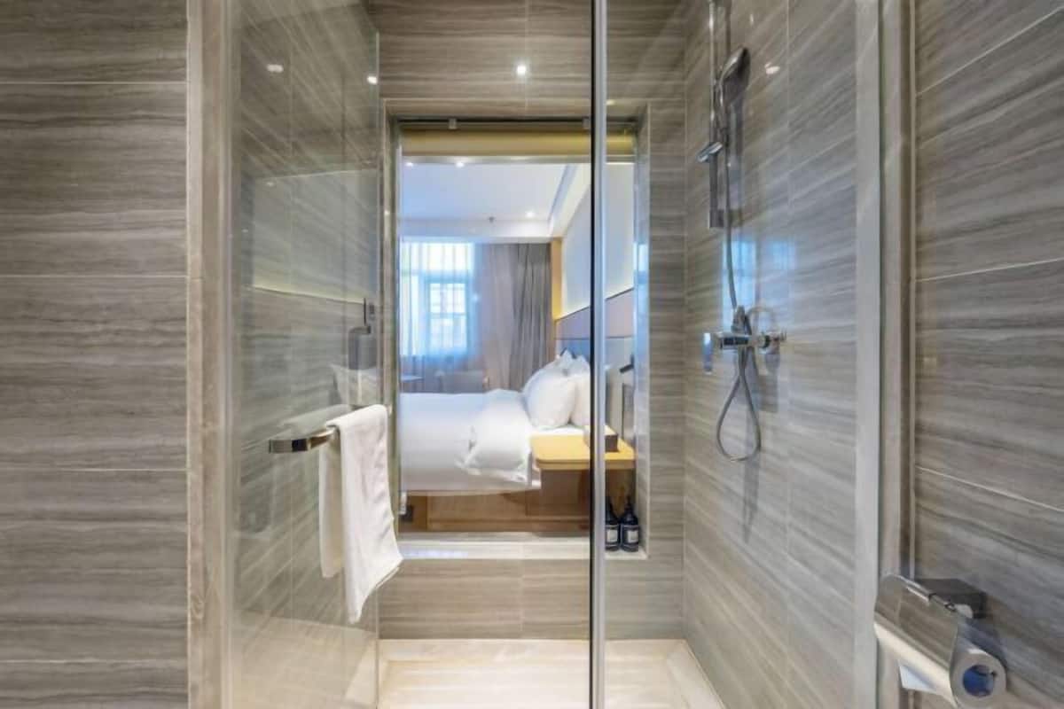 A modern bathroom with a glass partition, a shower, and a visible bedroom area with a bed and a nightstand.